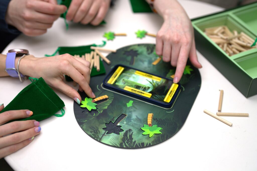 Pavaizduoti stalo žaidimo elementai: žaidimo lentos centre- mobilusis įrenginys su įjungta mobiliąja aplikacija, į lentą įsistatančios palmių figūrėlės, maišeliai ir pagaliukai, nerimstantys rankų sūkuryje 