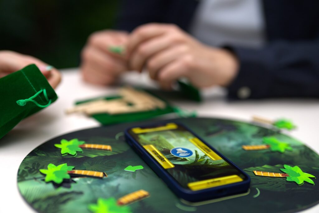 Iš arti pavaizduotas lentos centre esantis mobilusis įrenginys su įjungta „Kondigno“ aplikacija. Ekraną braukiant pirštu į skirtingas puses, pasigirsta skirtingo pobūdžio užduotys.  